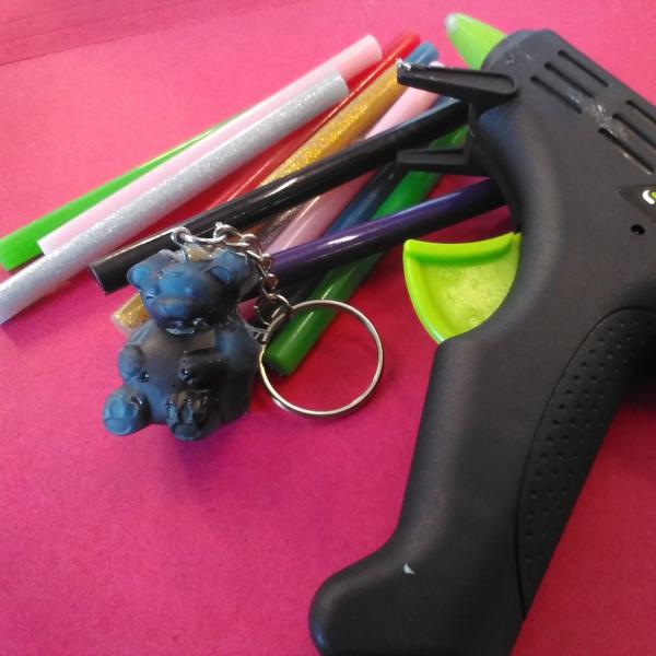 Multicolored hot glue sticks, a black and green hot glue gun, and a blue bear shaped hot glue keychain sit on a red background.
