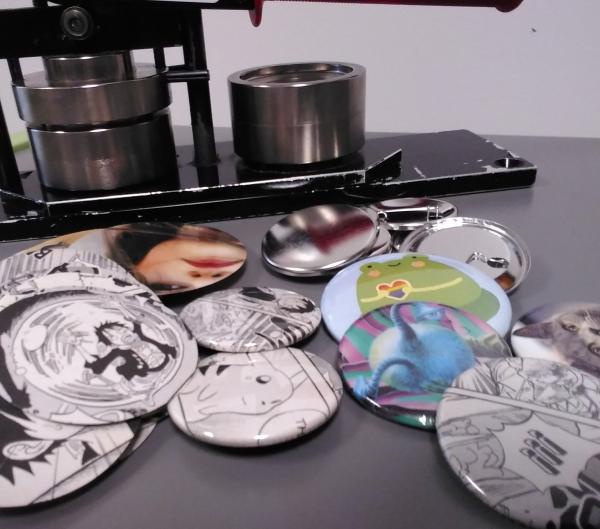 On a gray table, several circular paper clippings and buttons/badges are scattered in front of a button making press.