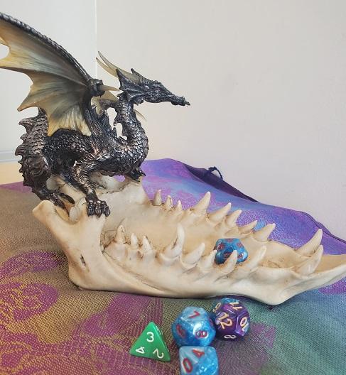 On a colorful surface, a green d4, a blue d8, a blue d12, and a purple d20 rest in the foreground. Behind them, a blue d20 rests inside a small statue/dice tray depicting a dark gray dragon sitting on the bottom jaw of an off-white animal skull.